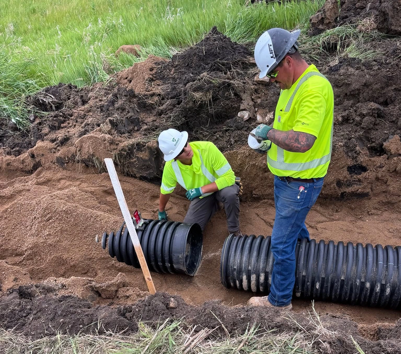 CM Student Anthony Albright crouches near piping during internship. Two people standing in dirt hole next to plastic piping