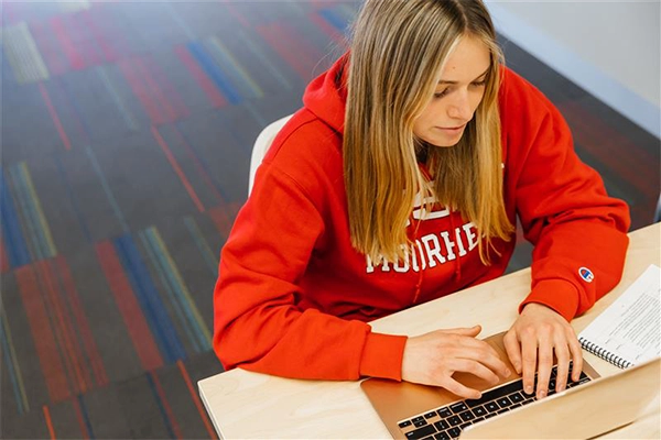 Student in red sweatshirt sitting at a table with a laptop